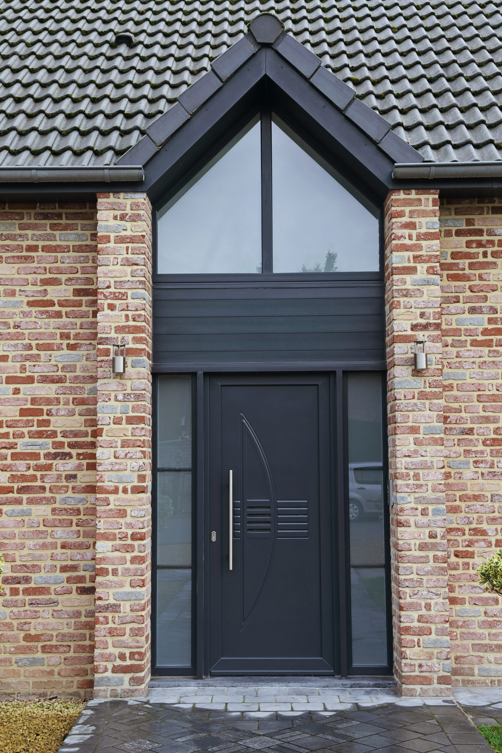 Porte d'entrée anthracite avec imposte vitrée triangulaire sur une façade en brique, réalisation Artisan du Châssis près de Mons