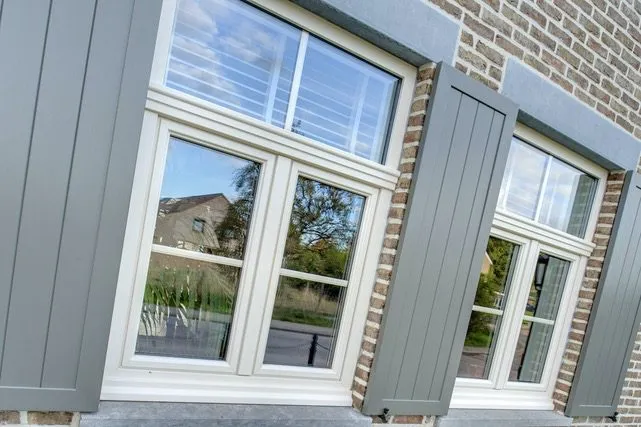 Fenêtres à frappe crème encadrées de volets battants gris sur une façade en brique, réalisation Sio2 Châssis à Flémalle