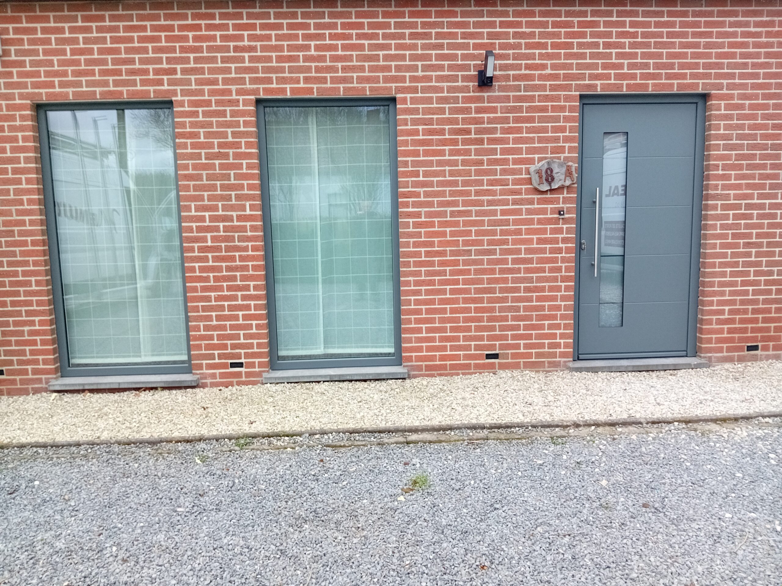 Deux châssis gris et porte d'entrée anthracite sur une façade en brique rouge, réalisation Menui'Deal dans la région de Mouscron