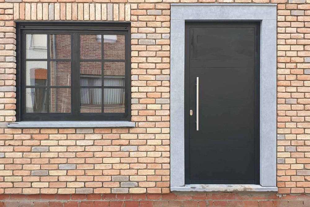 Fenêtre noire à petits-bois et porte d'entrée noire encadrées de pierre bleue sur une façade en brique, réalisation Menui'Deal près de Mouscron