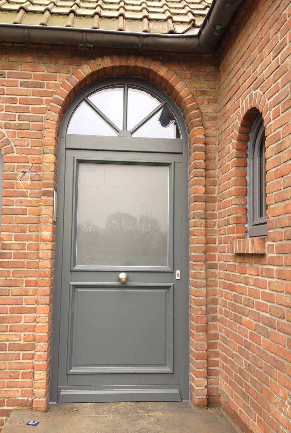 Porte d'entrée cintrée gris anthracite avec imposte vitrée sur une façade en brique, par Nikal Châssis