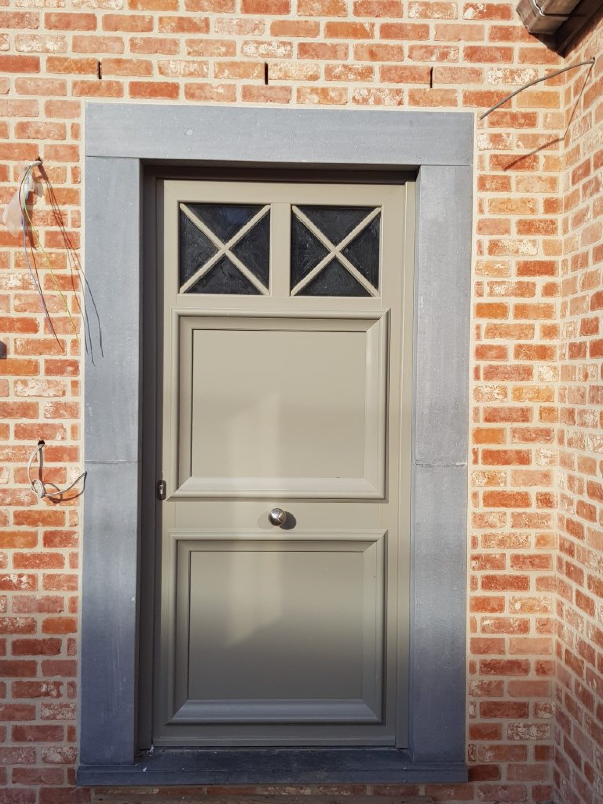Porte d'entrée taupe à imposte croisillonnée encadrée de pierre bleue sur une maison en brique, réalisation Chassis One à Wavre